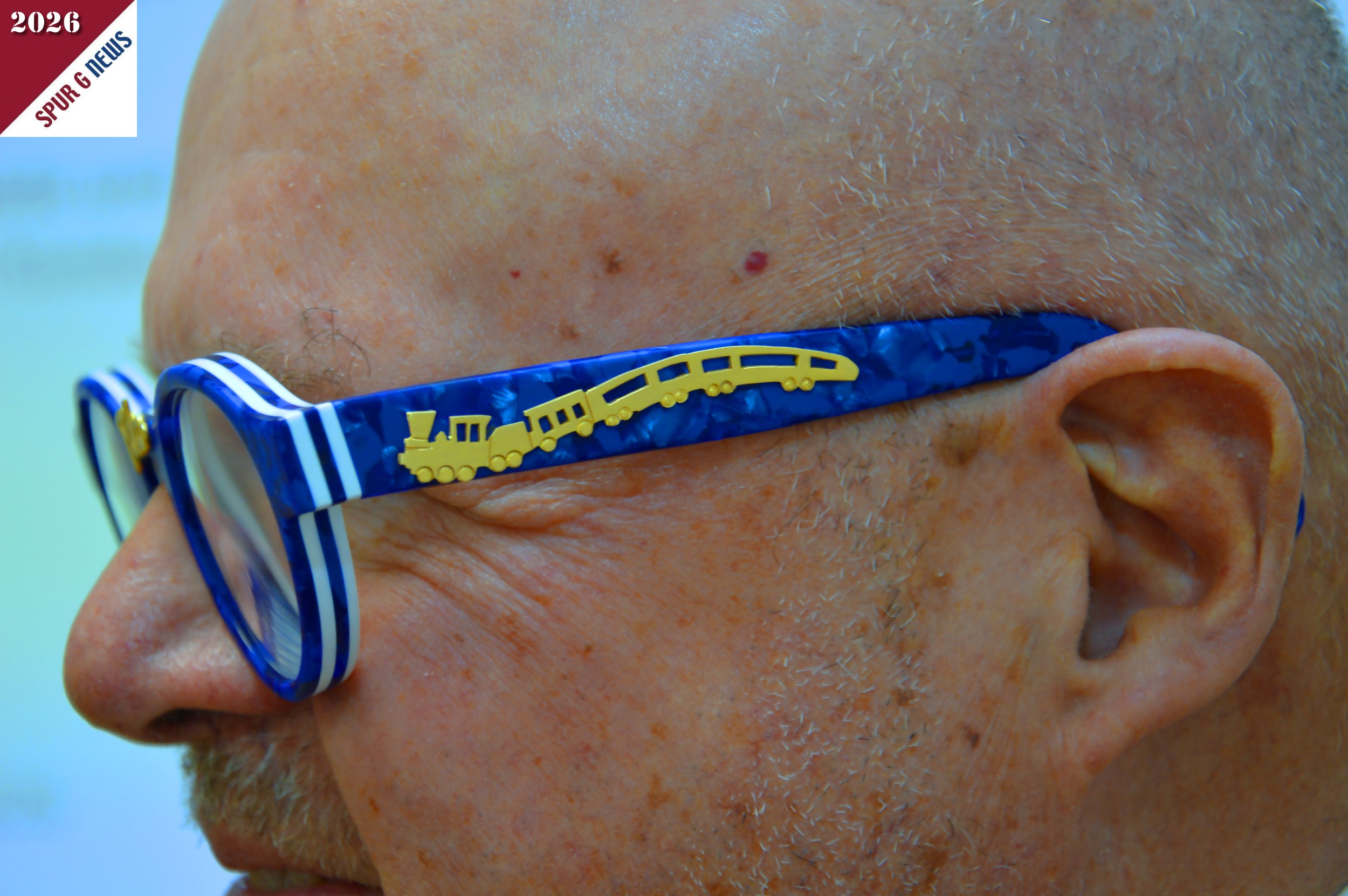 Auf dem Bild rechts ist die linke Seite der Brille von Herrn Dr. René F. Wilfer aus 2026 zu sehen. Auf dem Bügel ist ein geschwungene Darstellung eines Zuges von der Dampflok zum ICE zu sehen.