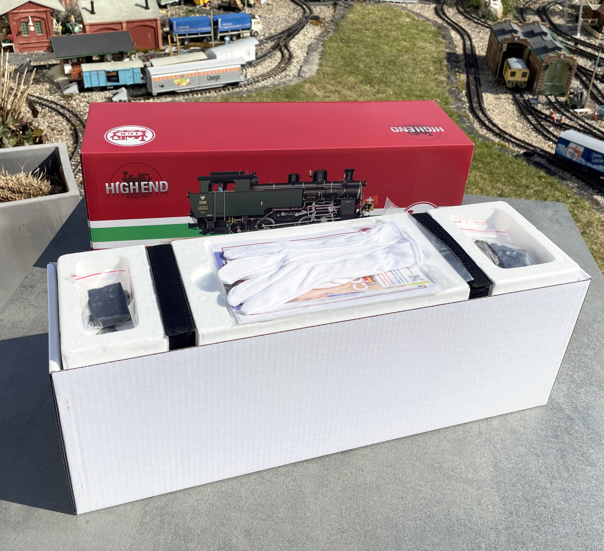 Der obere Teil des zweischaligen Verpackungskartons - rotes Oberteil - wei�es Innenteil - gibt den Blick auf die feste Styroporverpackung frei. Wer schon mal eine Highend Metalllok, z.B. Krokodil ausgepackt hat, kennt den Anblick mit den beiden Klettb�ndern. Links und rechts sind in den Kartonvertiefungen neben Magneten, Maulschl�ssel, Fahnen auch noch Kupplungen untergebracht. Im Mittelteil liegen die Baumwollhandschuhe, zur Vermeidung von Fingerabdr�cken auf der Lok, bei. Darunter das Zertifikat, der Achslenker f�r die Weichen, die Bedienungsanleitung und die Pipette zum Einf�llen den Dampflok�ls in den bereits werkseitig eingebauten Verdampfer.  